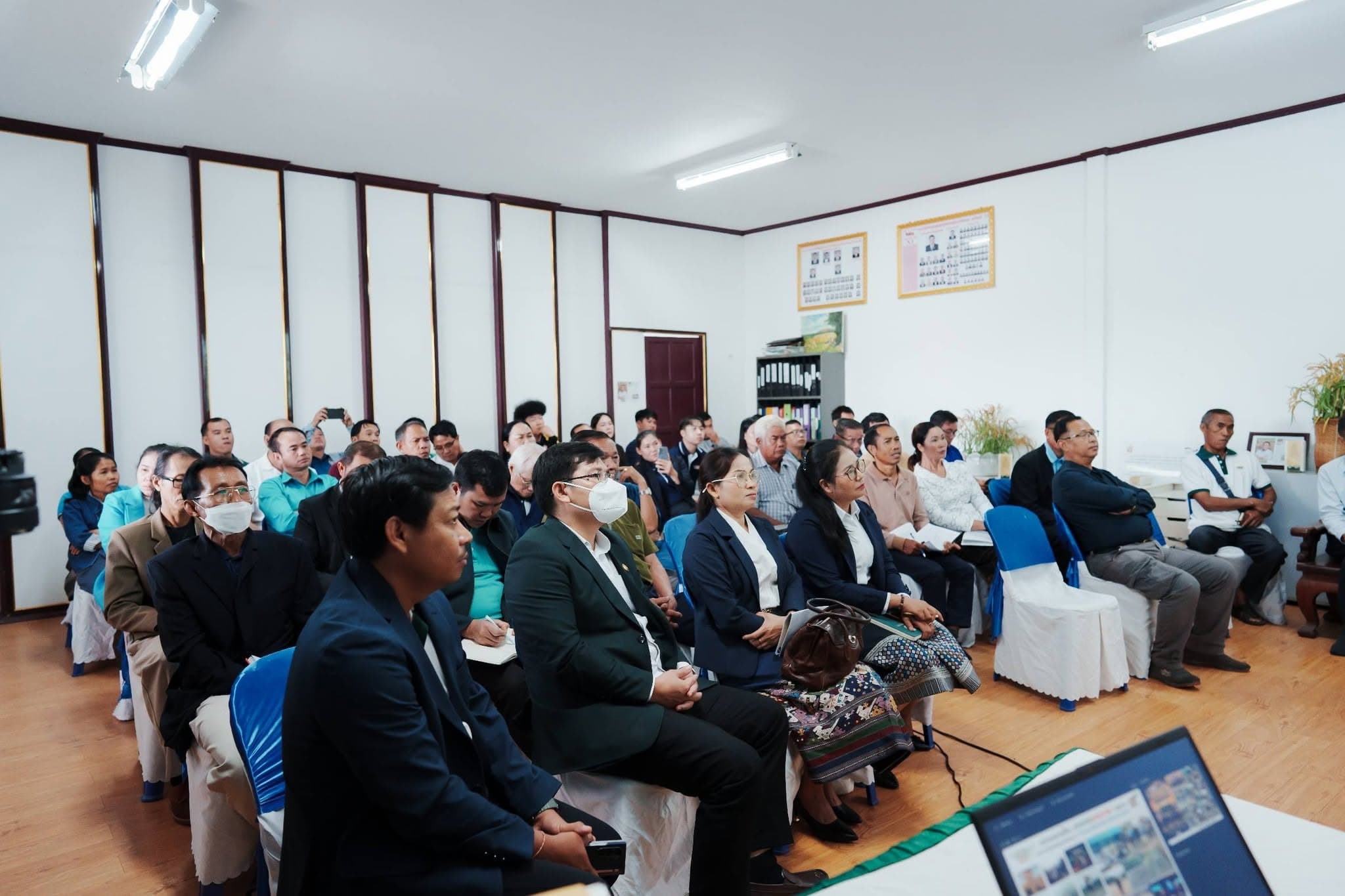 ผู้ฟังสวมสูทและผ้าซิ่นนั่งเรียงแถวในห้องประชุม หันหน้าไปทางวิทยากร