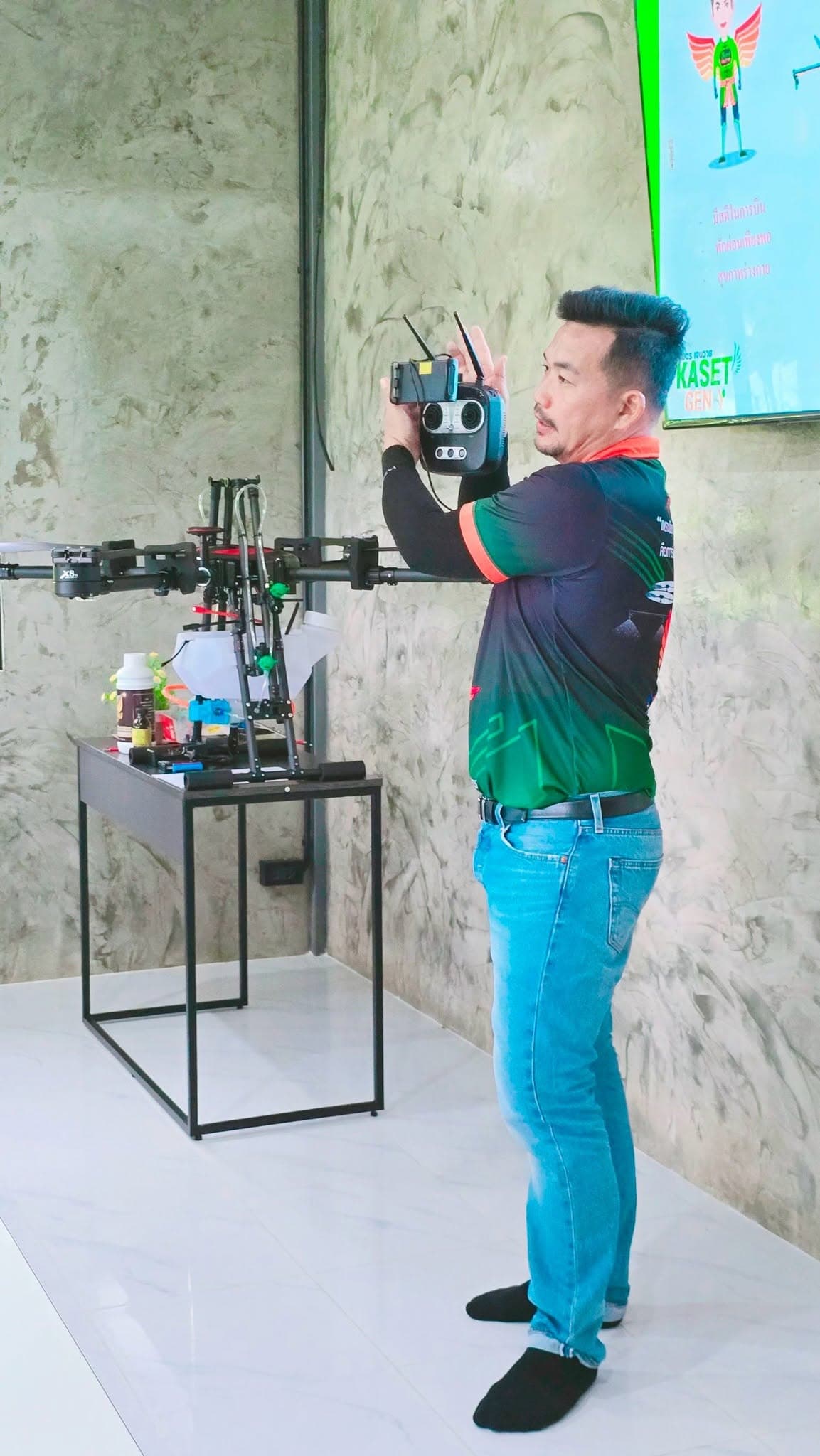 ครูฝึกยกรีโมตควบคุมขึ้นสาธิต ข้างโดรนที่ตั้งอยู่ในห้อง