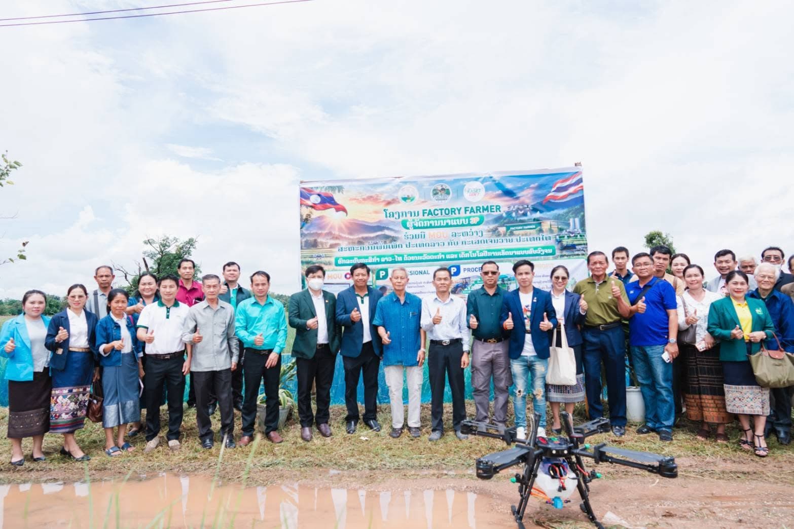 ผู้ร่วมงานยืนเรียงแถวริมแปลงนาหน้าป้ายโครงการ Factory Farmer ที่มีธงลาวและธงไทย โดรนพ่นยาวางอยู่บนพื้นด้านหน้า