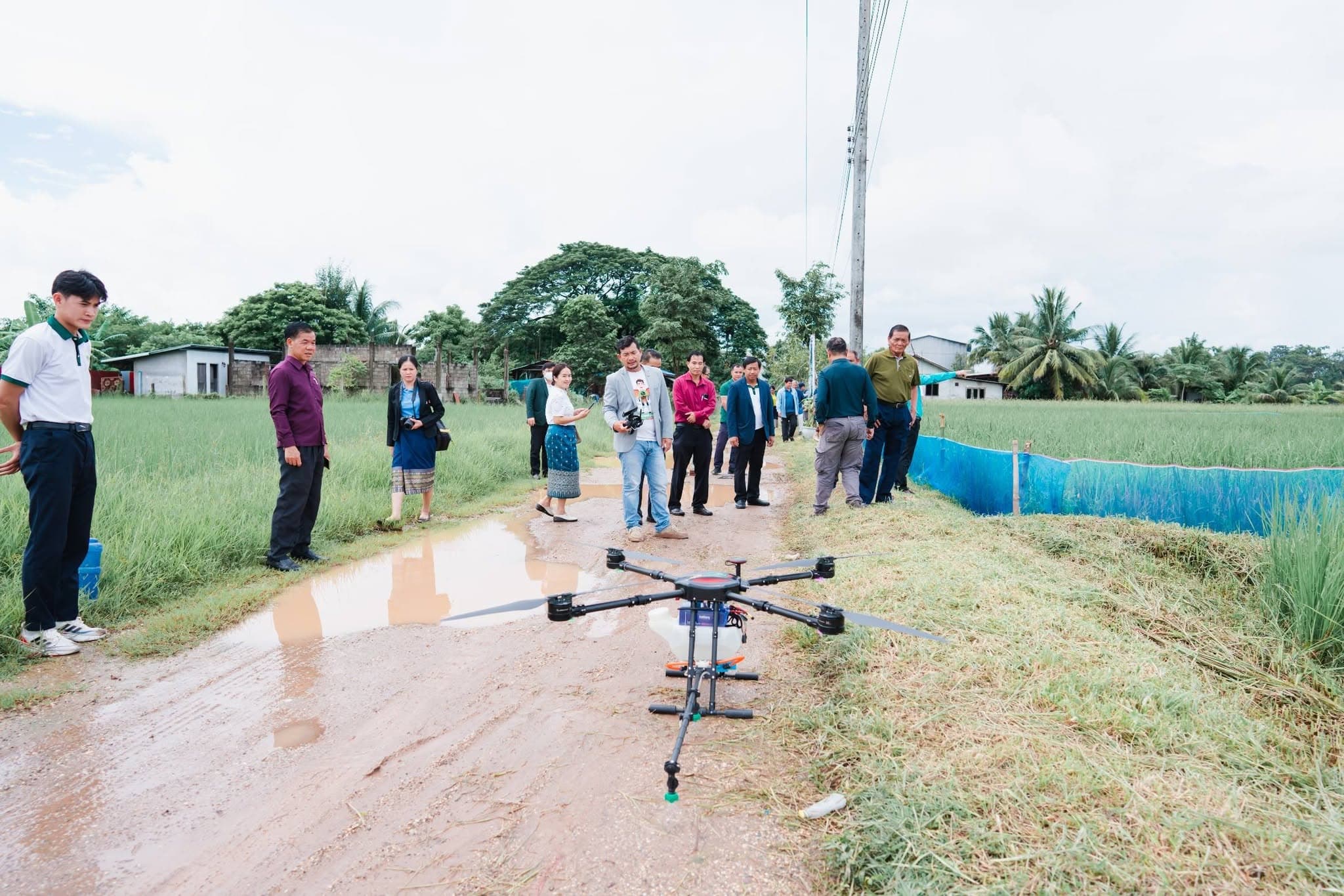 โดรนพ่นยาวางอยู่บนถนนดินข้างแปลงนา ผู้ร่วมงานยืนดูอยู่ด้านหลัง