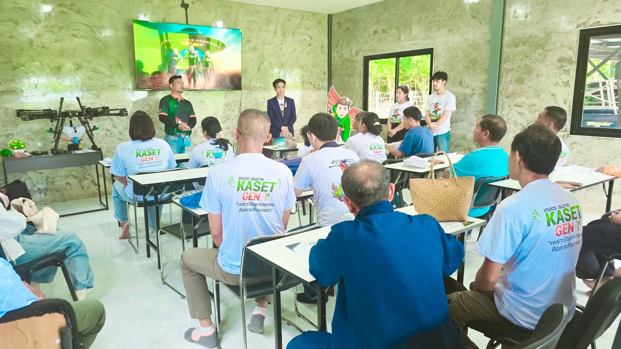 ผู้เข้าอบรมสวมเสื้อ KASET GEN-Y นั่งเรียนในห้องเรียน มีโดรนพ่นยาตั้งอยู่มุมห้อง