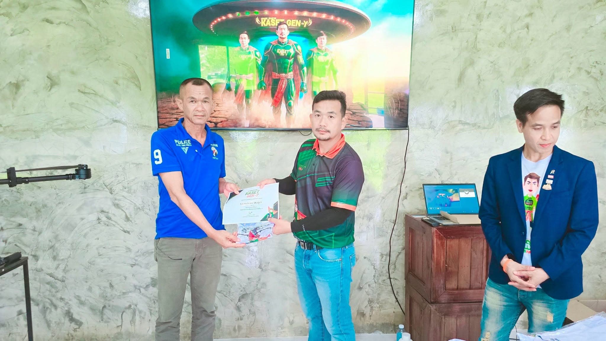 ผู้เข้าอบรมเสื้อสีน้ำเงินรับเกียรติบัตรจากครูฝึก วิทยากรสวมสูทยืนอยู่ข้าง ๆ