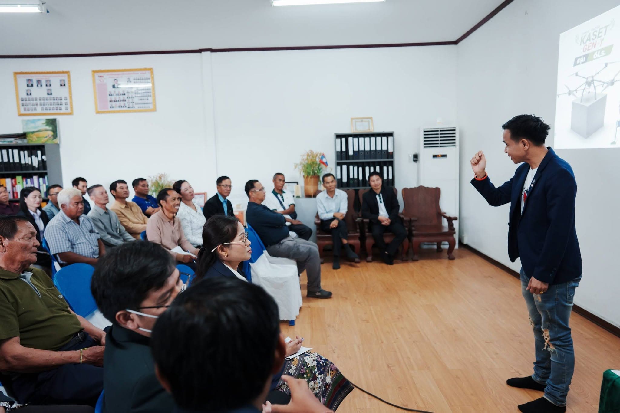 วิทยากรสวมสูทสีกรมท่ายืนพูดใกล้แถวผู้ฟัง ในห้องประชุมที่มีตู้แฟ้มเอกสาร