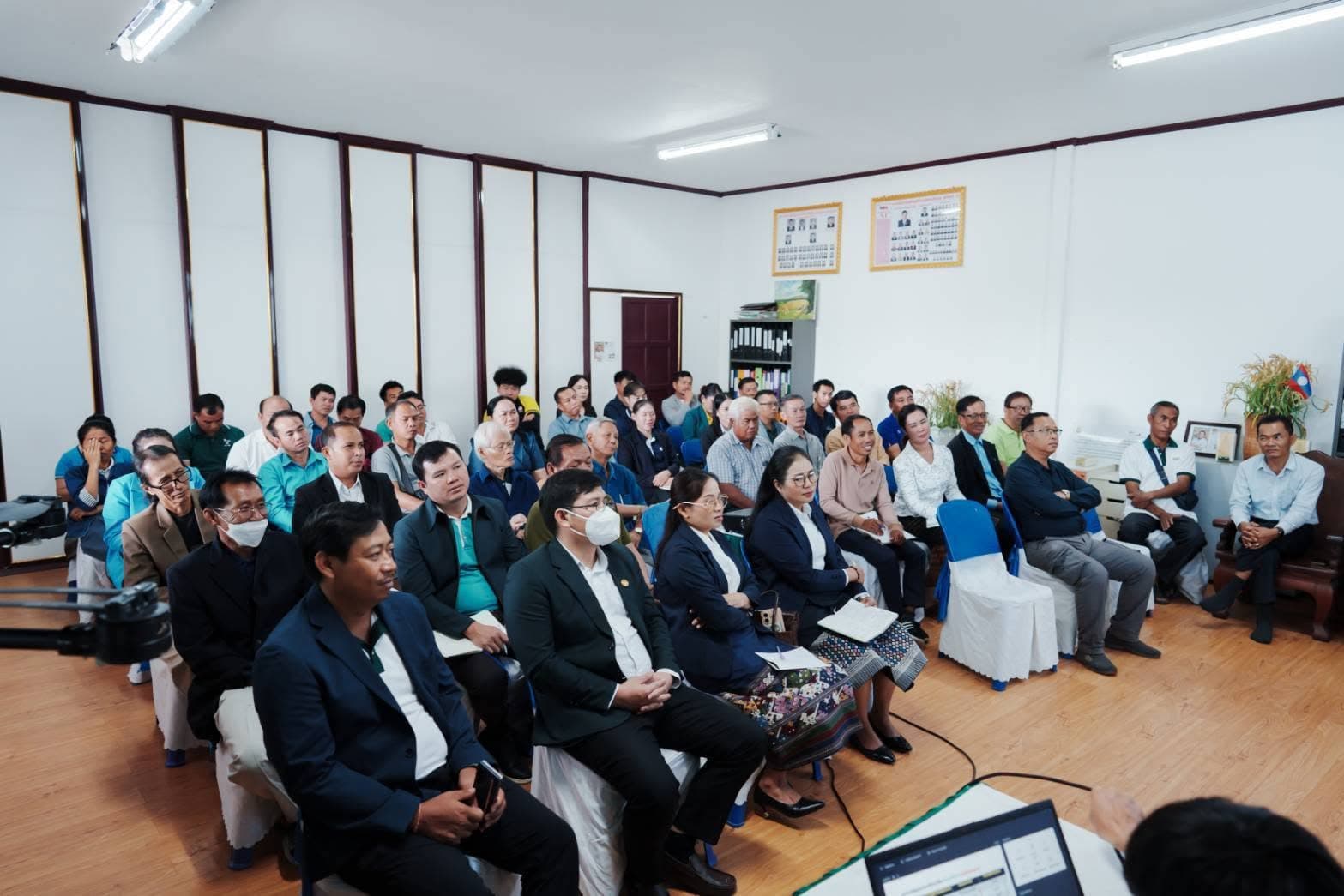 ผู้ฟังหลายสิบคนนั่งเรียงแถวในห้องประชุม บางคนสวมหน้ากากอนามัย