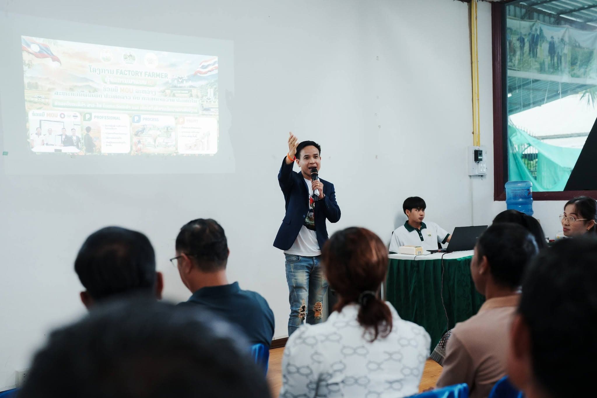 วิทยากรถือไมโครโฟนชี้ไปทางจอที่ฉายสไลด์โครงการ Factory Farmer ผู้ฟังนั่งด้านหน้า